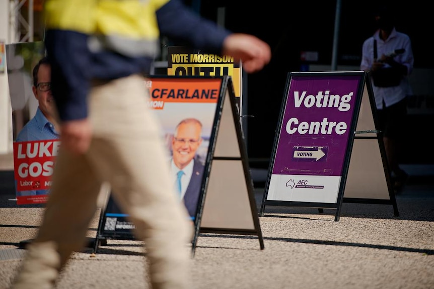 A man in high-vis shirt walks past a collection of corflutes on sandwich boards and a sign saying 'Voting Centre' with an arrow