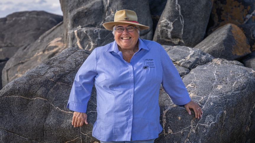 A woman in a blue, long-sleeved shirt stands against large rocks.