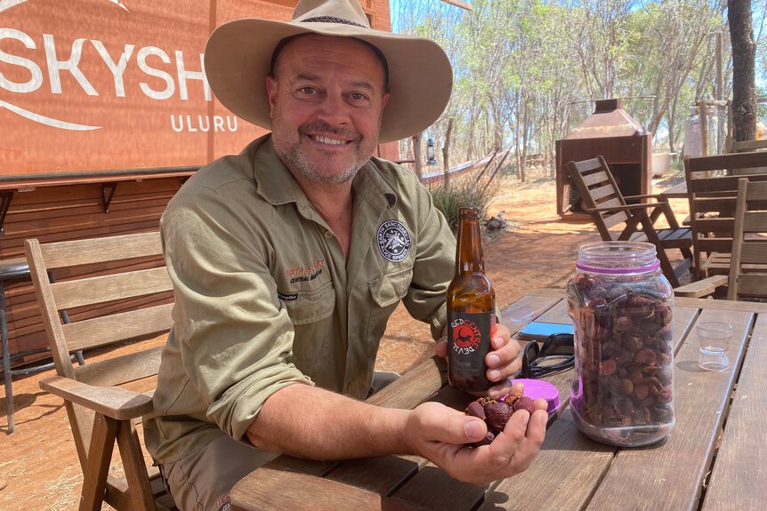 A man wearing a khaki work uniform and hat holds a handful of dried purple, pink quandongs and a beer with a red label