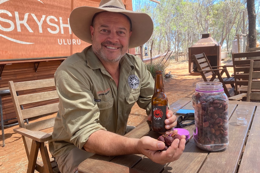 A man wearing a khaki work uniform and hat holds a handful of dried purple, pink quandongs and a beer with a red label