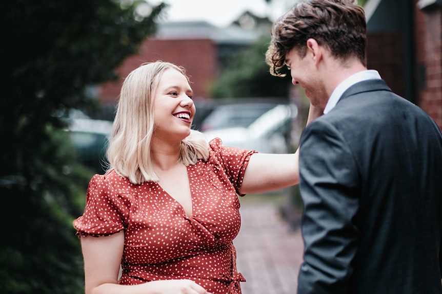 A woman with a red spotty dress smiles at a man standing next to her. He's wearing a suit.