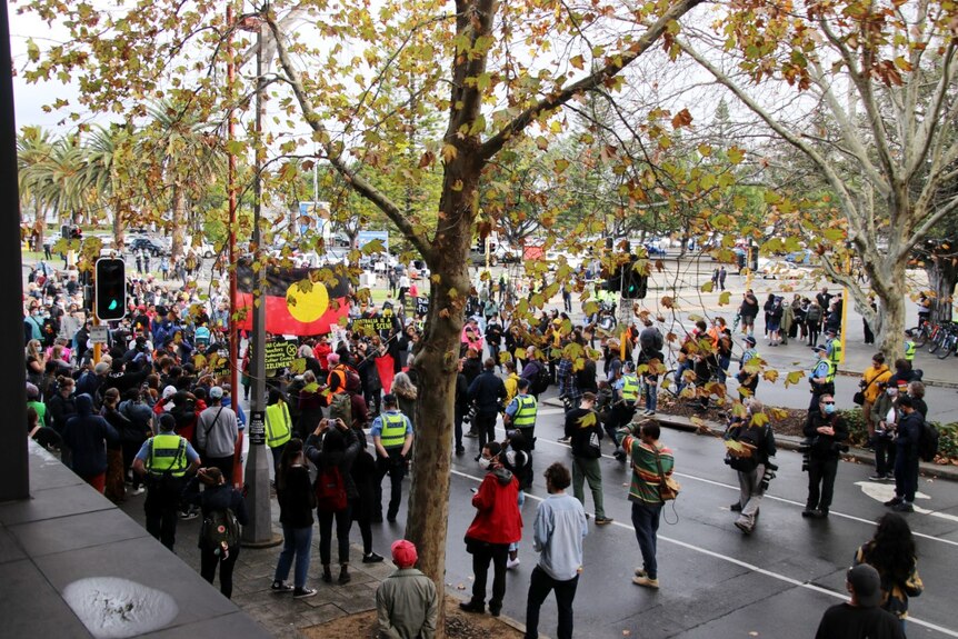 A crowd of people stand at a Perth intersection in front of trees with a large Aboriginal flag displayed.