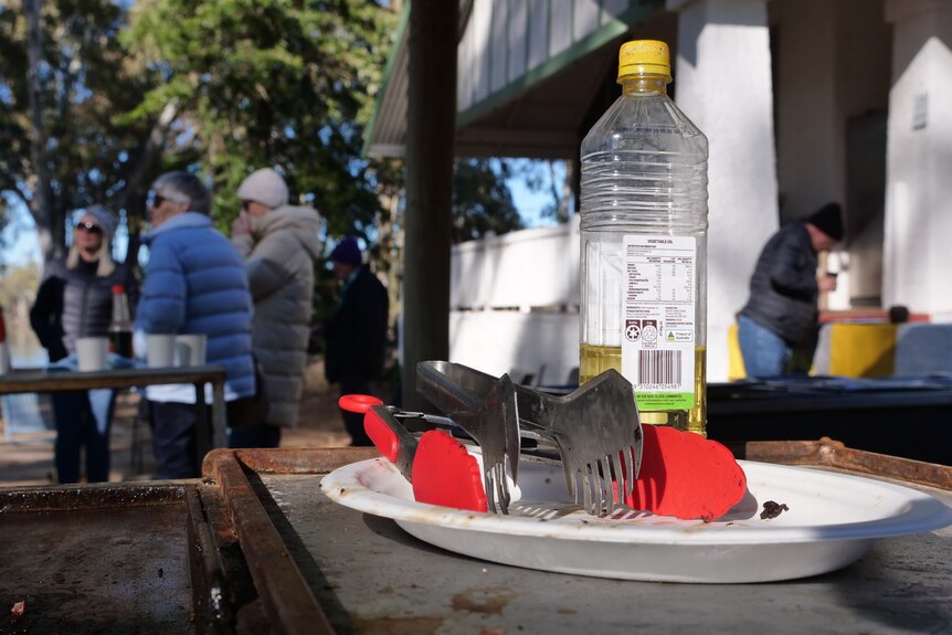 A close up of tongs and oil for the barbeque bacon and eggs. People are gathered in the background.