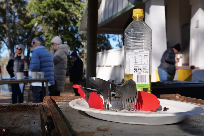 A close up of tongs and oil for the barbeque bacon and eggs. People are gathered in the background.