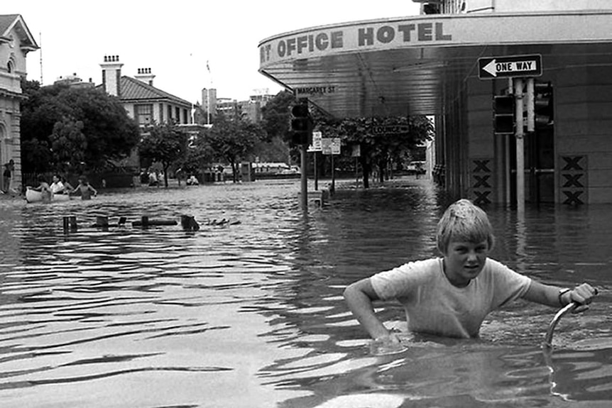 A black and white photo of a boy pushing his bike through chest-high floodwater.