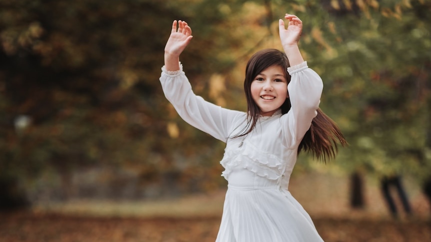 Happy girl in a flowing white dress is twirling under an autumn tree 