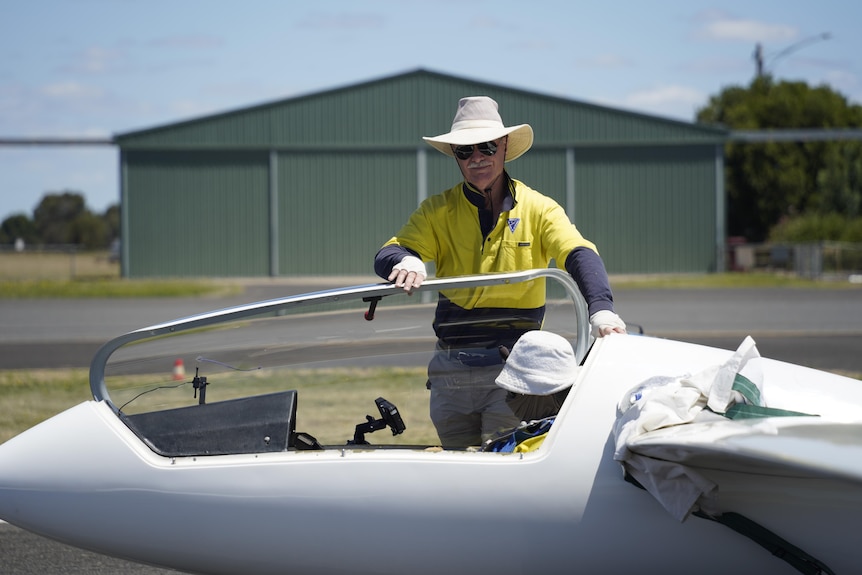 A man and a woman prepare to take-off in a large white glider, both wear white hats, yellow shirts.