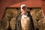 A farm in farming overalls and a checked jacket stands in front of a barn full of circular hay bales.