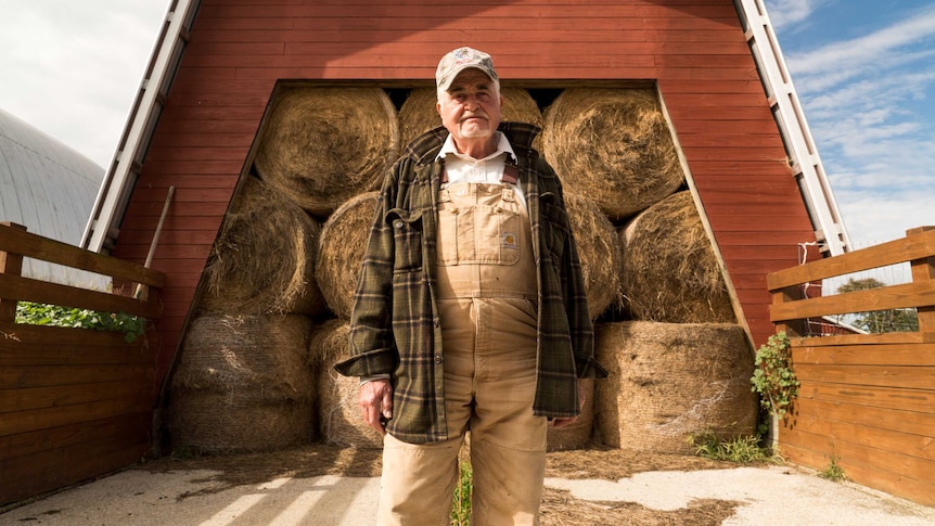 A farm in farming overalls and a checked jacket stands in front of a barn full of circular hay bales.