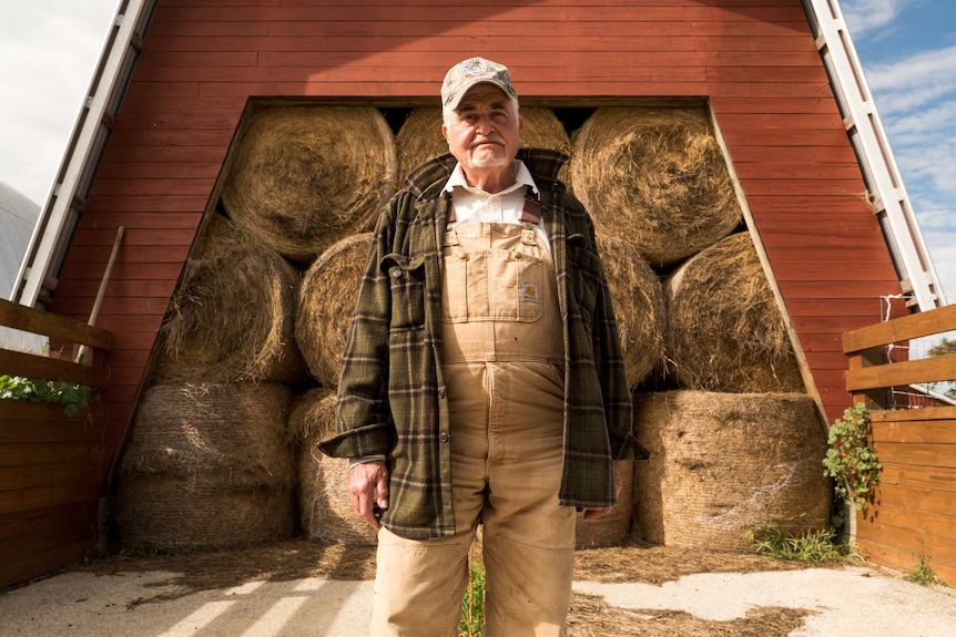 A farm in farming overalls and a checked jacket stands in front of a barn full of circular hay bales.