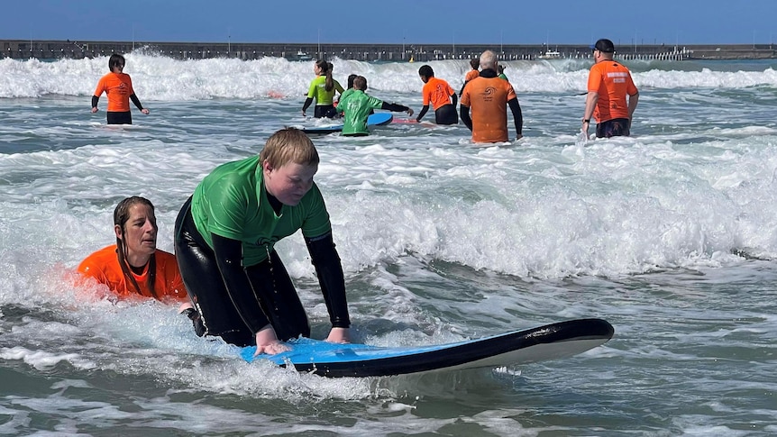 A boy kneels on a beginner surfboard with help from woman in water behind