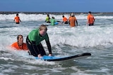 A boy kneels on a beginner surfboard with help from woman in water behind