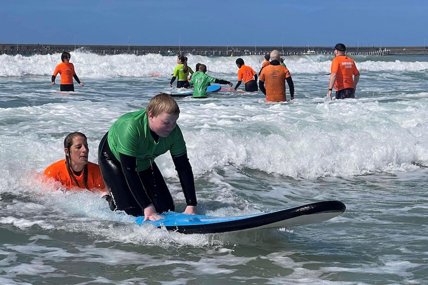 A boy kneels on a beginner surfboard with help from woman in water behind