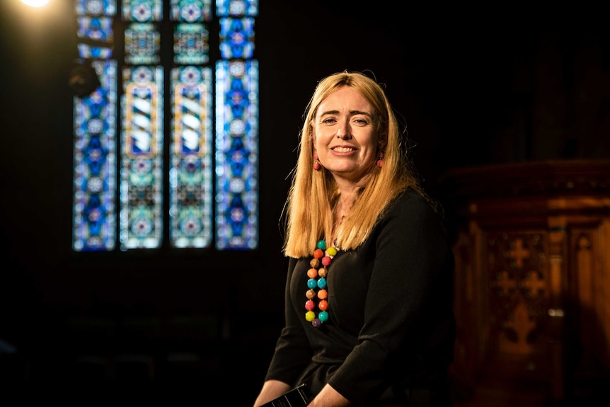 Rev Dr Dani Treweek sits inside a church with a stained glass window behind her.