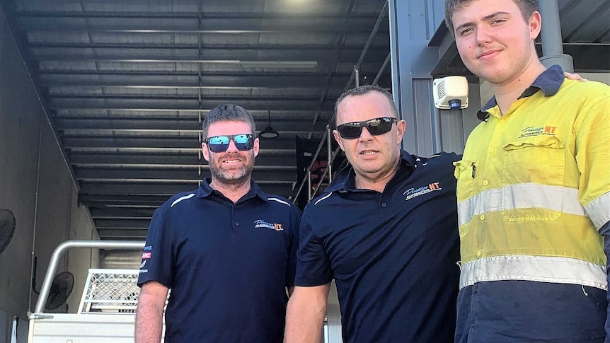 Three auto-electricians wearing company attire and sunglasses standing outside their workshop.