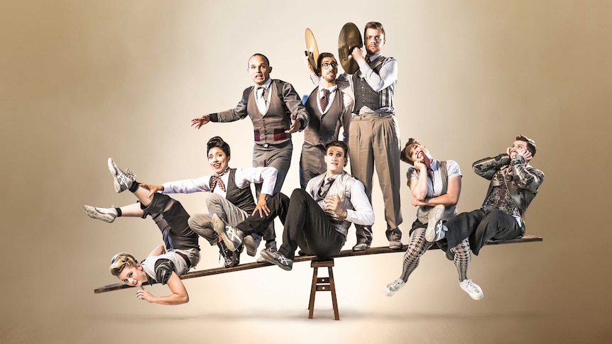 Eight circus performers wearing suits balance on a see-saw in front of a beige backdrop.