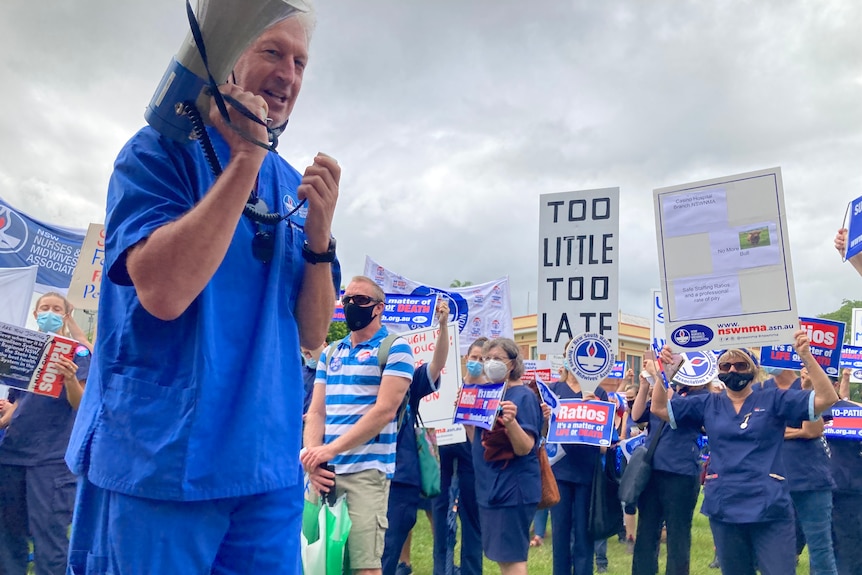 Man wearing blue scrubs holding loud speaker. Crowd holding signs behind him 