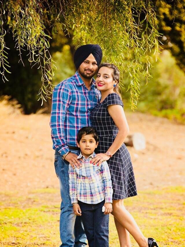 A man wearing a Sikh turban stands with his wife and son underneath a green leafy tree smiling at the camera.