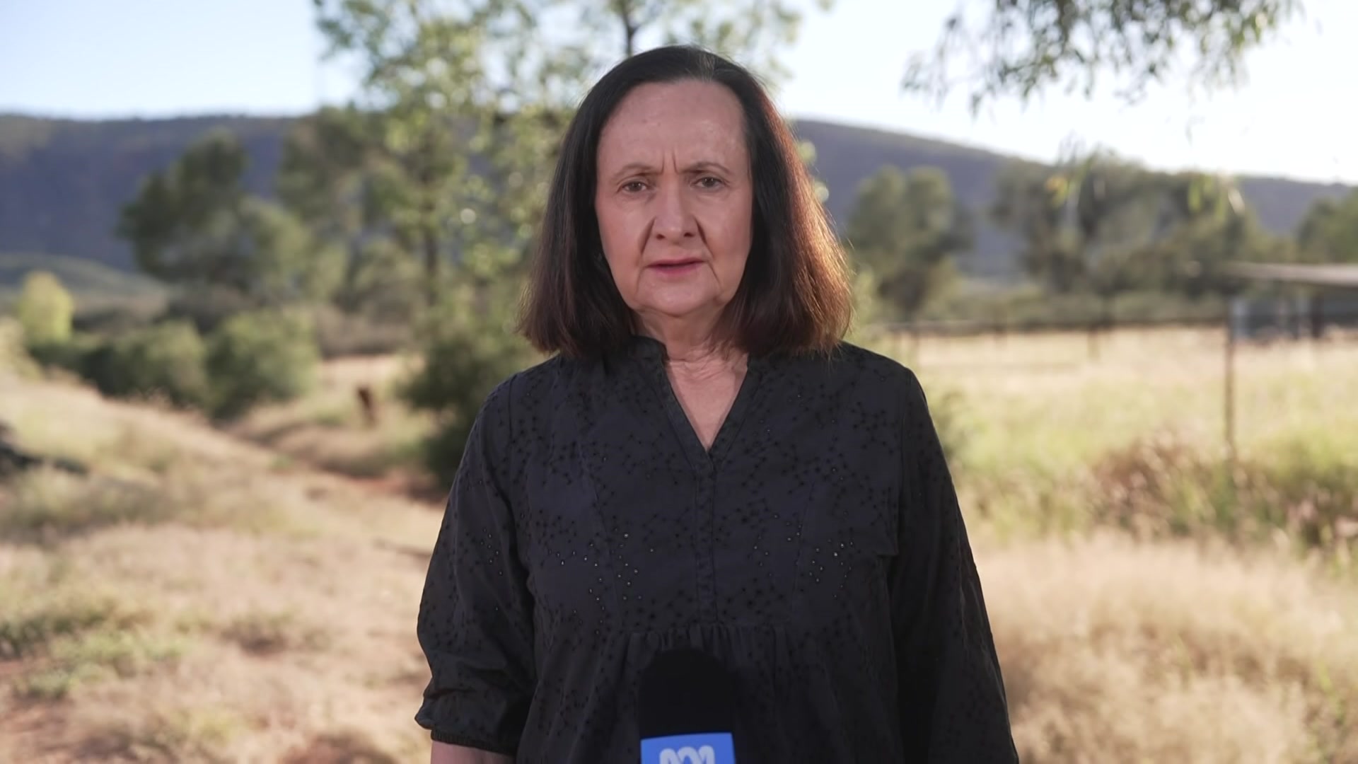 A woman standing in a sparse scrubland landscape, holding a microphone and talking to the camera.