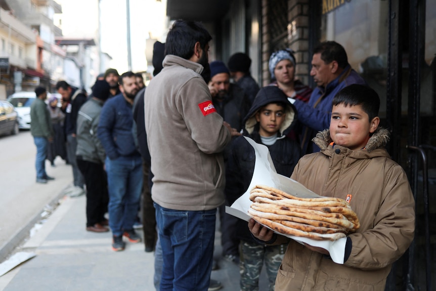 A boy holds a pile of bread as people line up behind him. 