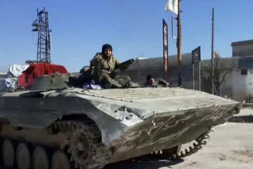 A man sits on top of a tank which is rolling through a city.