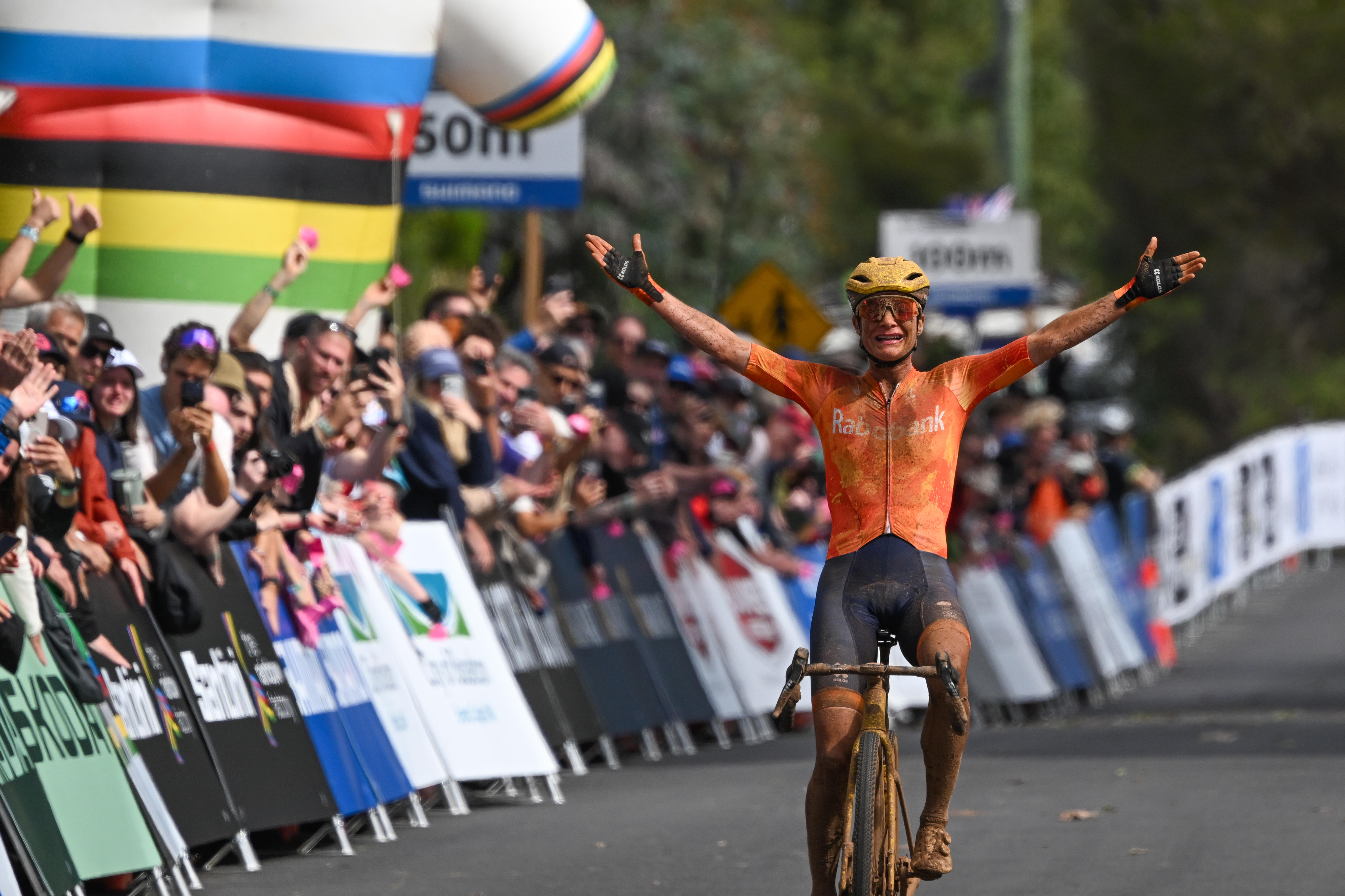 A cyclist wearing orange and back has arms outstretched.