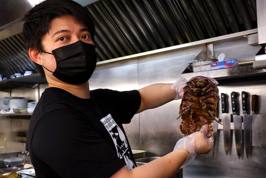 A Chinese man with black mask and black T-shirt holds a crab like creature in kitchen