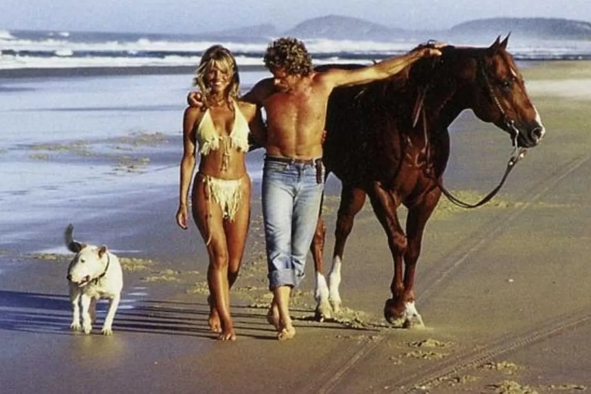 Alby Mangels and a bikini-clad Judy Green walk on the beach with a horse and a dog.