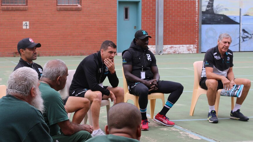 Three port adelaide footballers face us as prisoners face them listening to them talk. Tall red brick walls all around