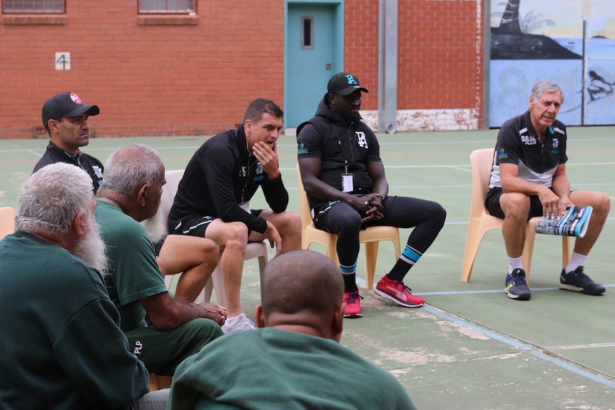 Three port adelaide footballers face us as prisoners face them listening to them talk. Tall red brick walls all around