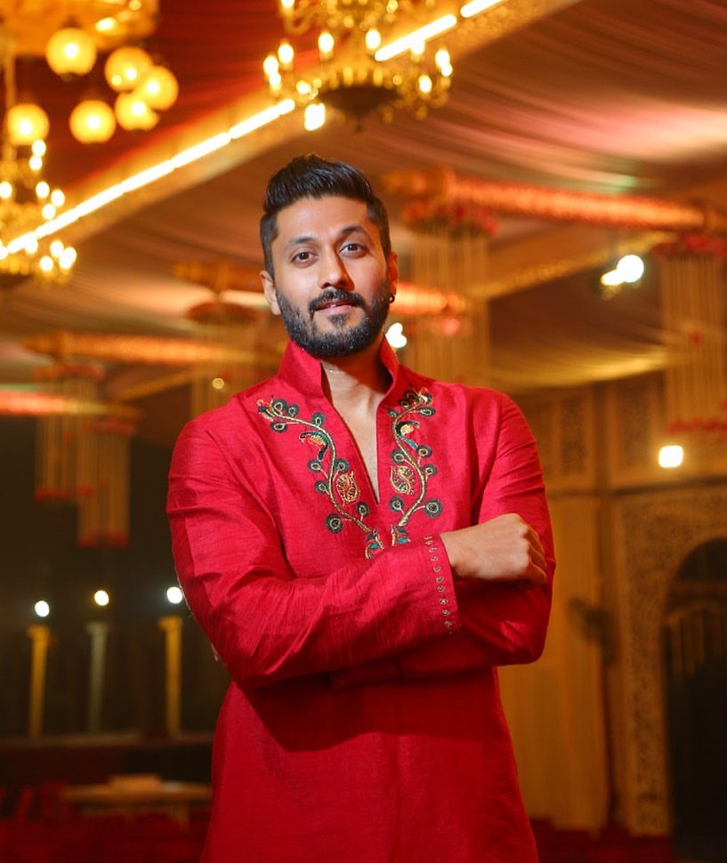Chetan Kumar Ahimsa wearing a red shirt staring at the camera with his arms folded