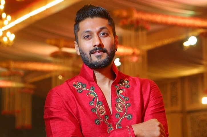 Chetan Kumar Ahimsa wearing a red shirt staring at the camera with his arms folded