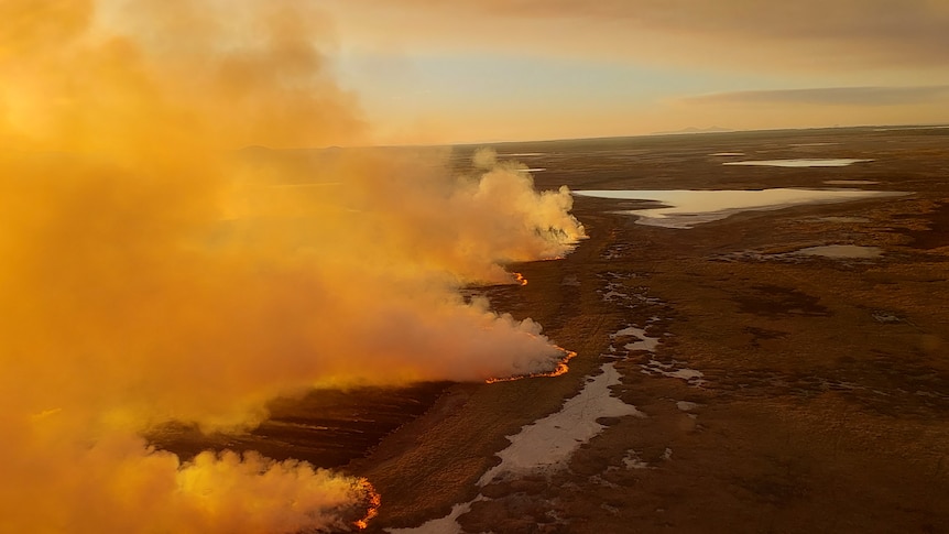 An aerial view of an orange smokey fire on land, sea in the distance.