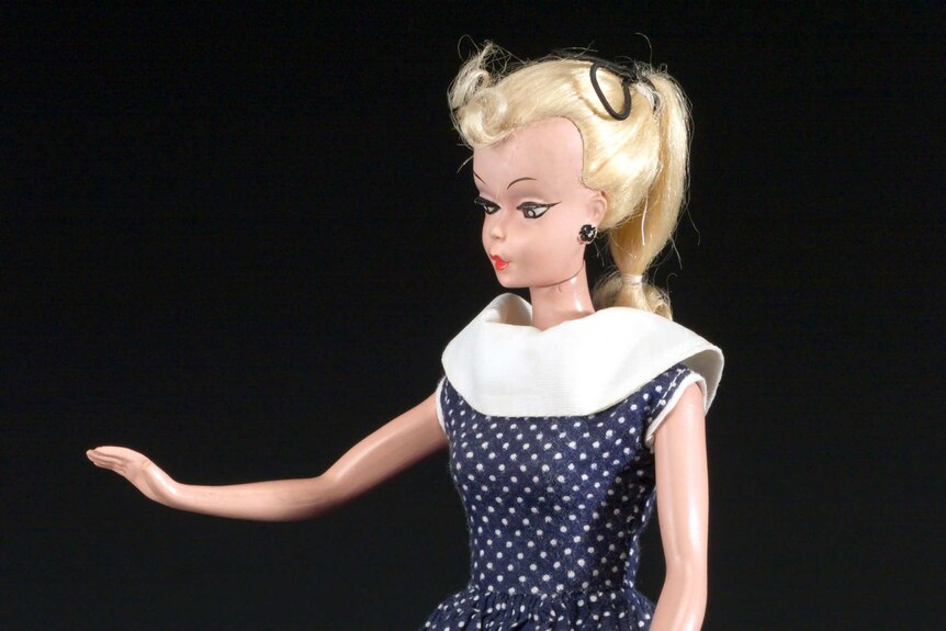 A plastic doll with rigid limbs sits, wearing a polka-dot dress, black high heel shoes and make-up, and with blonde hair.