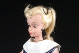 A plastic doll with rigid limbs sits, wearing a polka-dot dress, black high heel shoes and make-up, and with blonde hair.