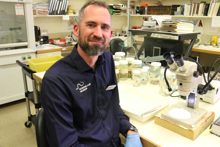 A man in a navy shirt sits next to a microscope smiling