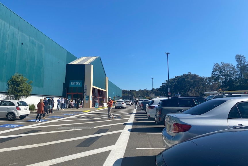 Dozens of cars parked outside a large warehouse and a long line of people lined up at the entry.