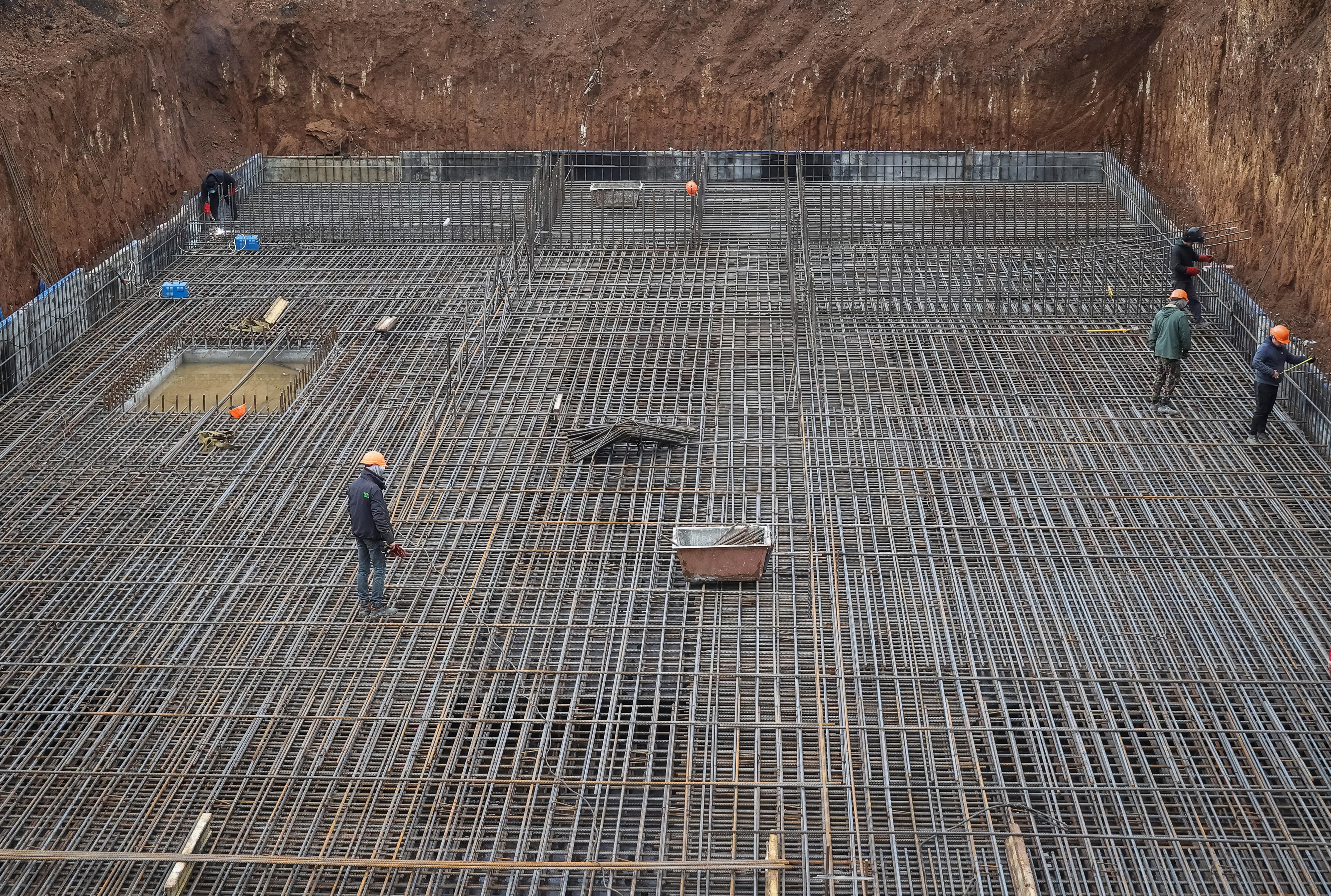 Men wearing hardhats are pictured from afar on a metal structure, around it is a building site.