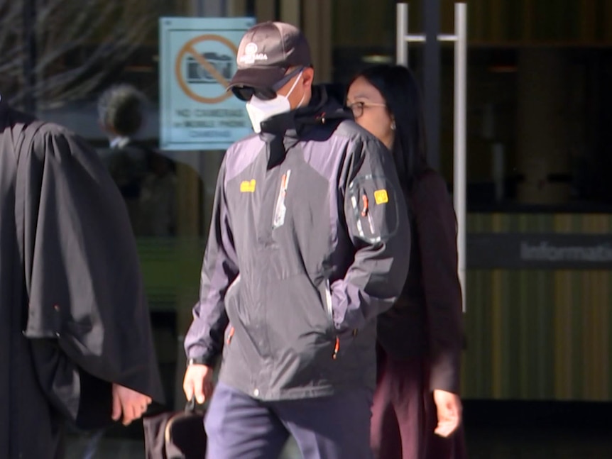 A man in a black windbreaker, black cap, black sunglasses and white face mask walks next to a man in a barrister's gown.