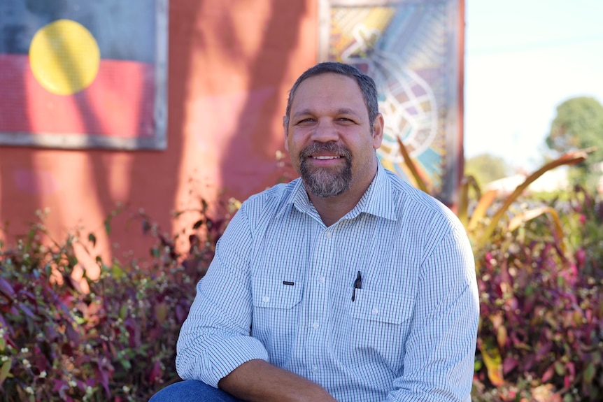 A man looks at the camera and smiles. He's crouching in front of a mural with Aboriginal flag painted on it.