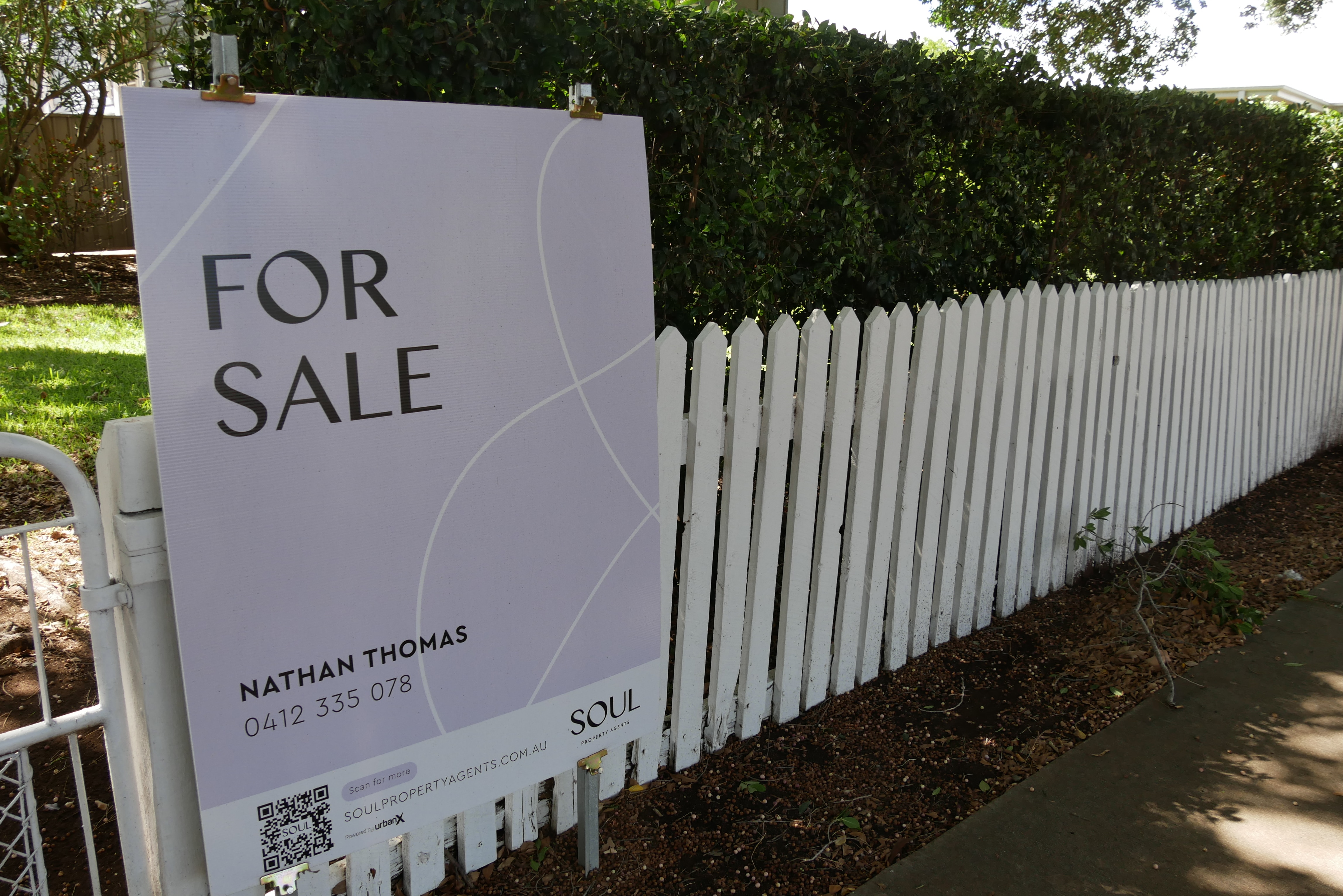 Un cartel de venta de color púrpura se asienta sobre una valla blanca con un seto verde detrás. 