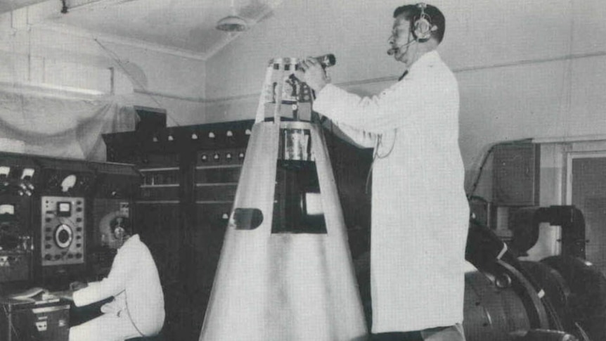 A black and white photo of a man in a lab coat standing on a bench working on the top of a rocket tip.