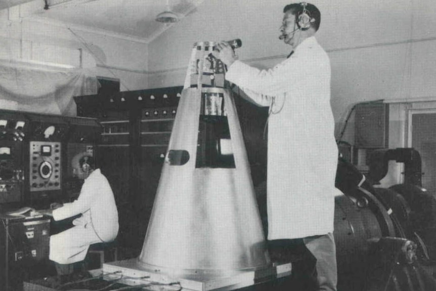 A black and white photo of a man in a lab coat standing on a bench working on the top of a rocket tip.