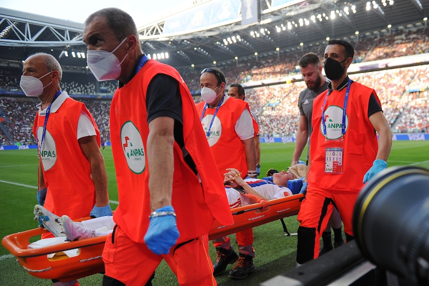 Australian footballer Ellie Carpenter lies on a stretcher being carried off the field by four men.