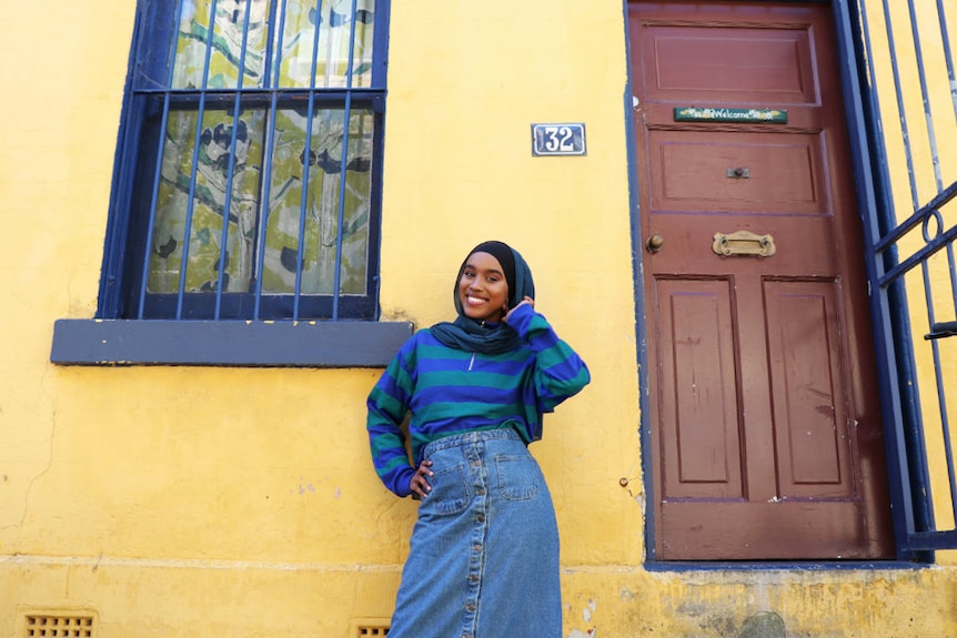 Hijabi model Miski Omar poses in front of bright yellow house to depict modest fashion.