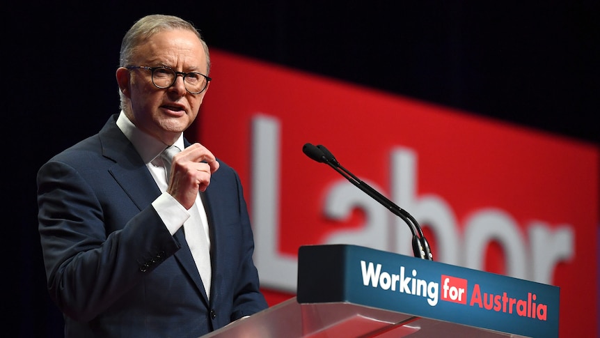 A man wearing glasses and a suit stands behind a lectern labelled "Working for Australia".