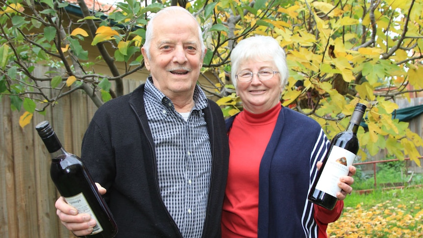 A man and woman stand in their garden, each holding a bottle of red wine.