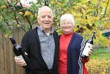 A man and woman stand in their garden, each holding a bottle of red wine.