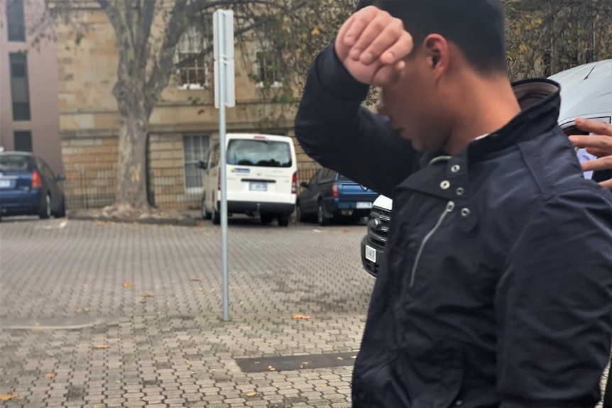 A man wearing a navy jacket walking along the street with his arm covering his face.