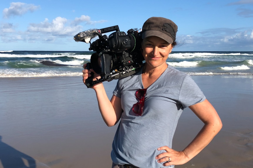 Picture of the filmmaker wearing light tee and dark cap, with camera on shoulder, at the beach, surf in background.